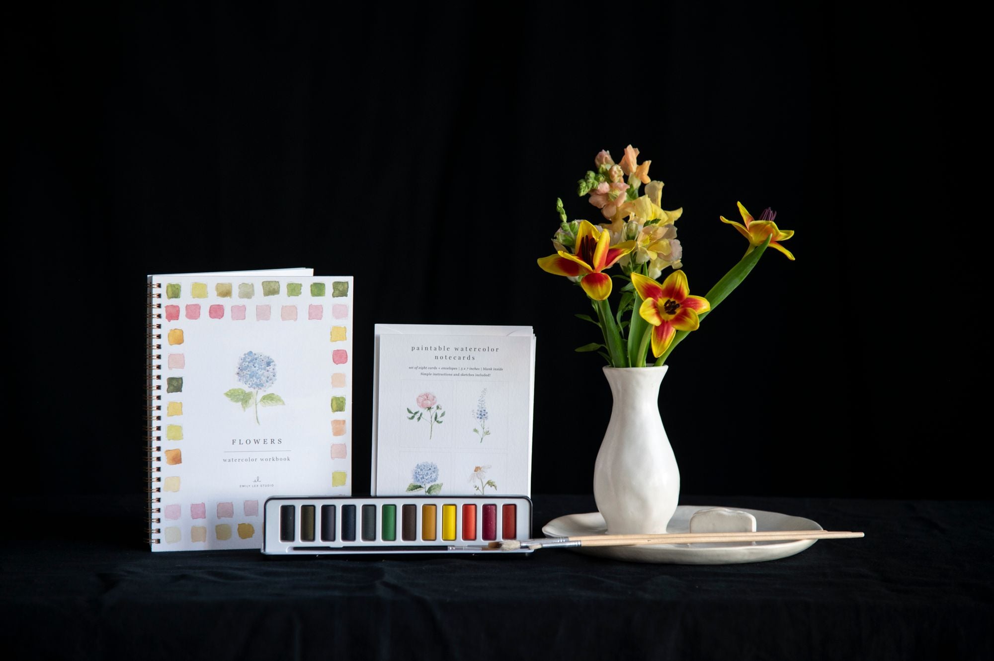 Art supplies including a notebook, watercolor palette, and flowers in a vase