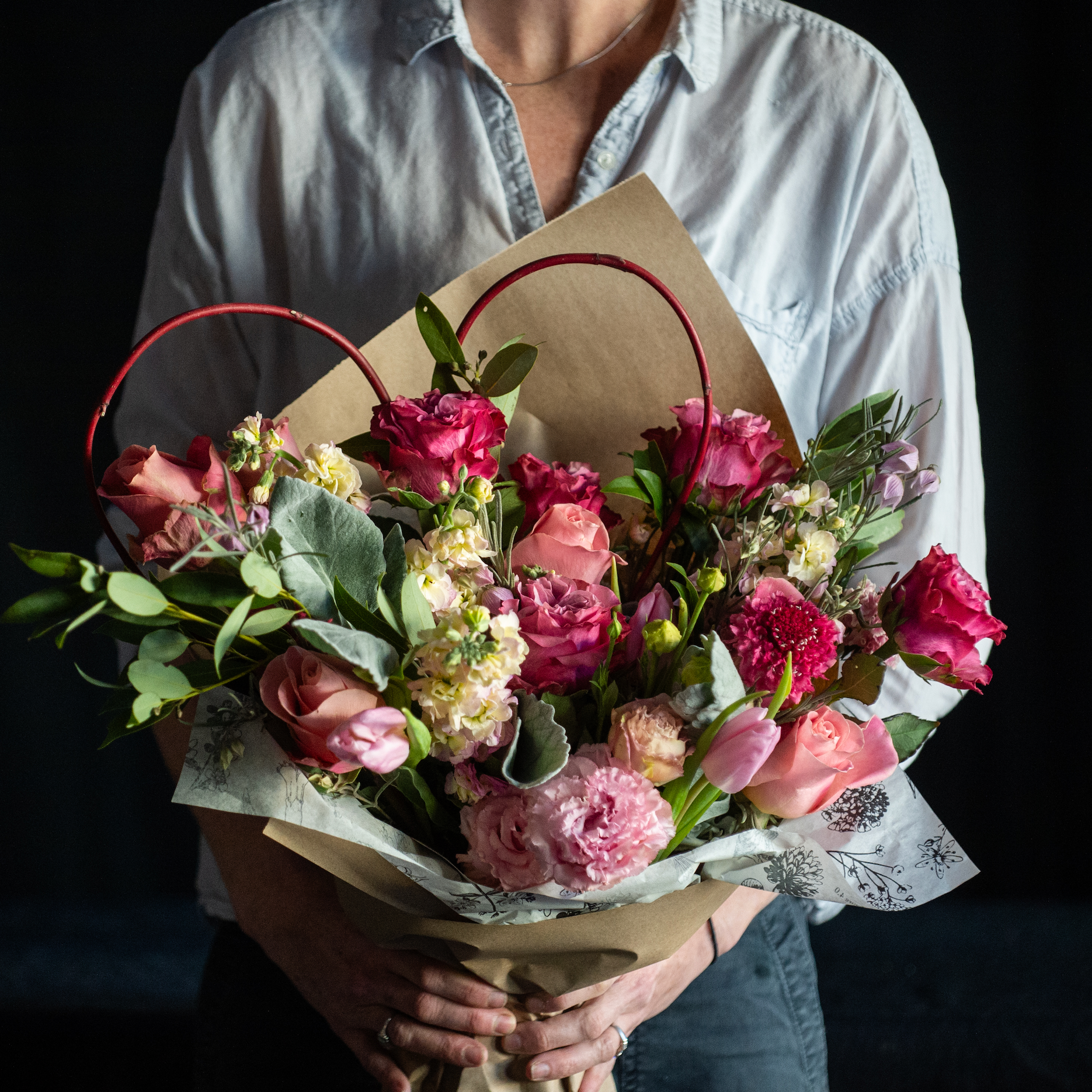 A wrapped bouquet of pink roses with premium accent stems with a red branch heart.