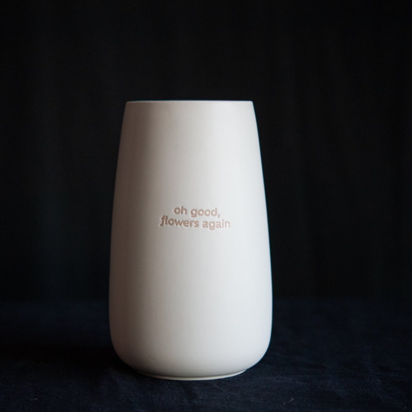 A white ceramic vase that has the message "oh good, flowers again"