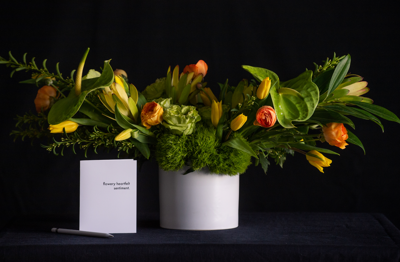 Modern horizontal floral design of green anthurium, green roses, tulips, ranunculus, and premium greenery. Next to a standard greeting card for scale