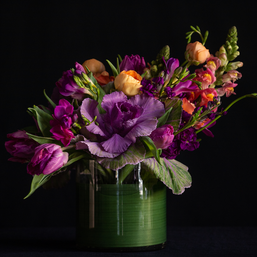 An asymmetrical design of kale, freesia, ranunculus, and snapdragons in a leaf lined container. Made by a local florist.