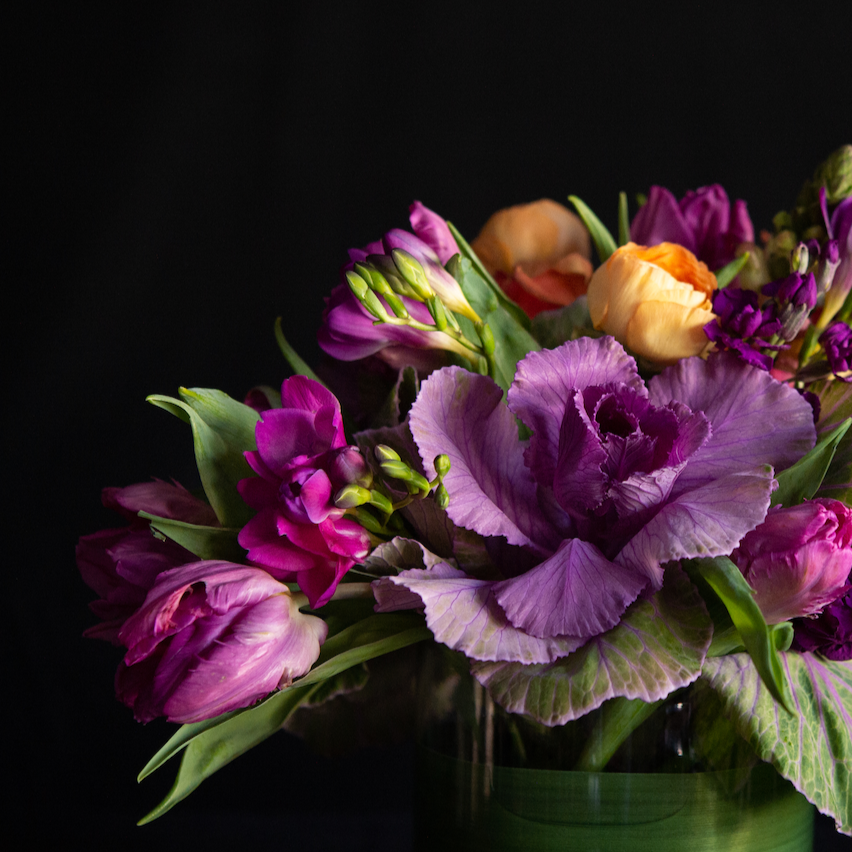 An asymmetrical design of kale, freesia, ranunculus, and snapdragons in a leaf lined container. Made by a local florist.