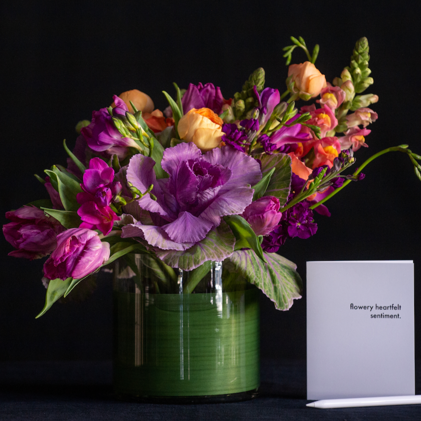 An asymmetrical design of kale, freesia, ranunculus, and snapdragons in a leaf lined container. Made by a local florist.