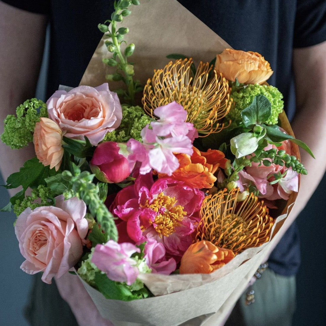 Wrapped bouquet of Spring flowers and peonies accented by tulips, hydrangea, roses, ranunculus, or tropicals plus greenery