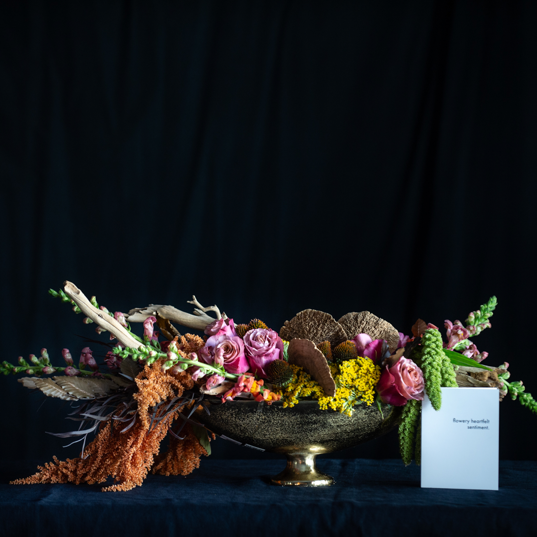 Autumn floral centerpiece in a bronze compote featuring roses, snapdragons, amaranthus, yellow yarrow, dried pods, and driftwood accents against a dark background.