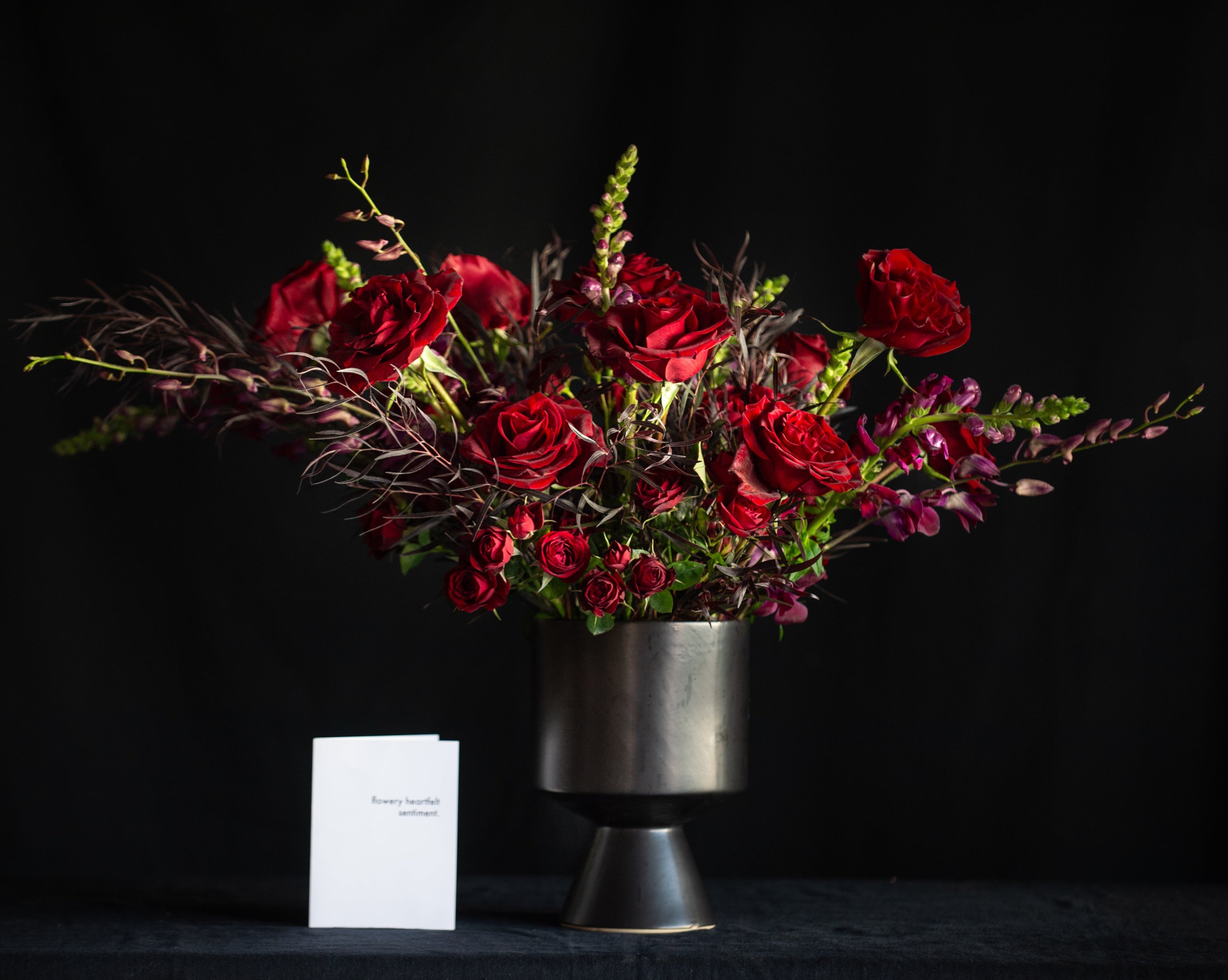 A dozen red roses with orchids, snapdragons, and dark foliage in a black footed vase