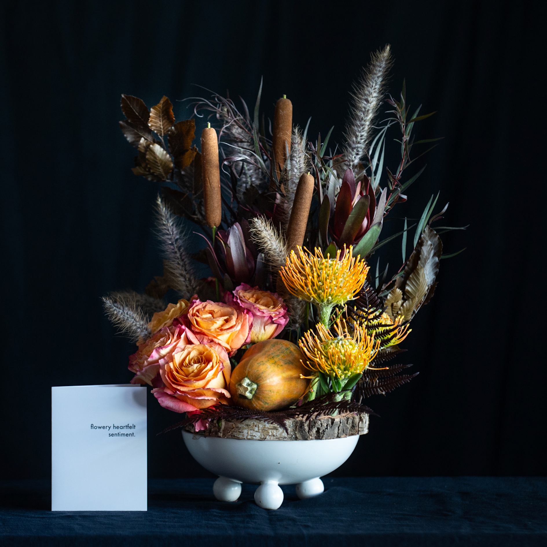 Fall floral arrangement with protea, roses, cattails, autumn foliage, and a small gourd in a white pedestal vase and wood charger.