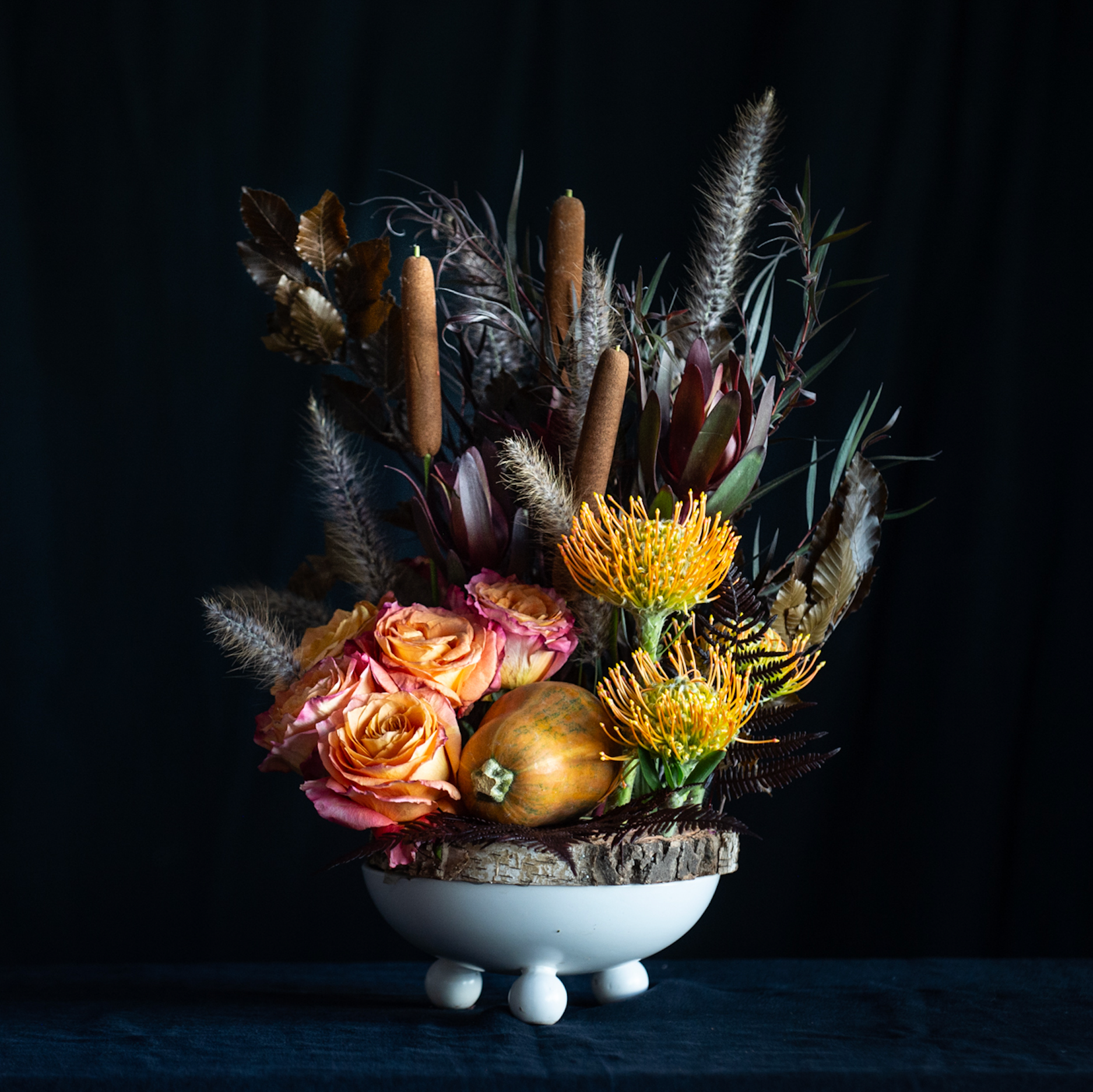 Fall floral arrangement with protea, roses, cattails, autumn foliage, and a small gourd in a white pedestal vase and wood charger.