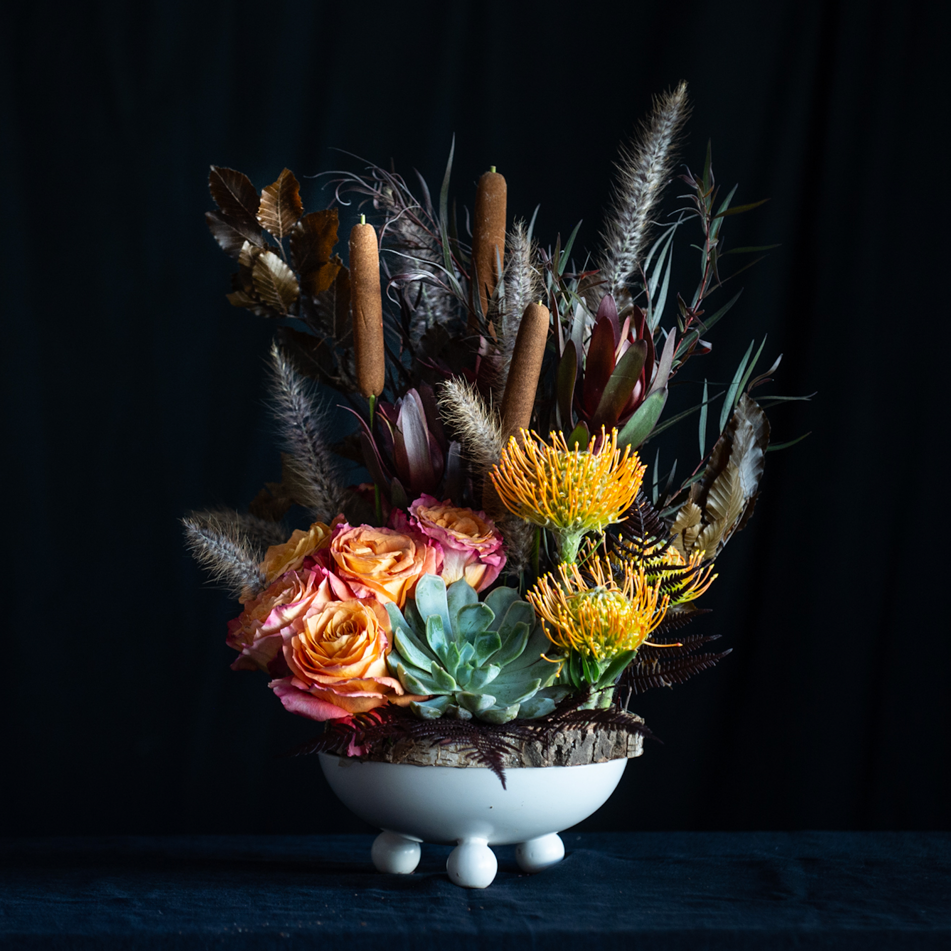 Fall floral arrangement with protea, roses, cattails, autumn foliage, and a succulent in a white pedestal vase and wood charger.