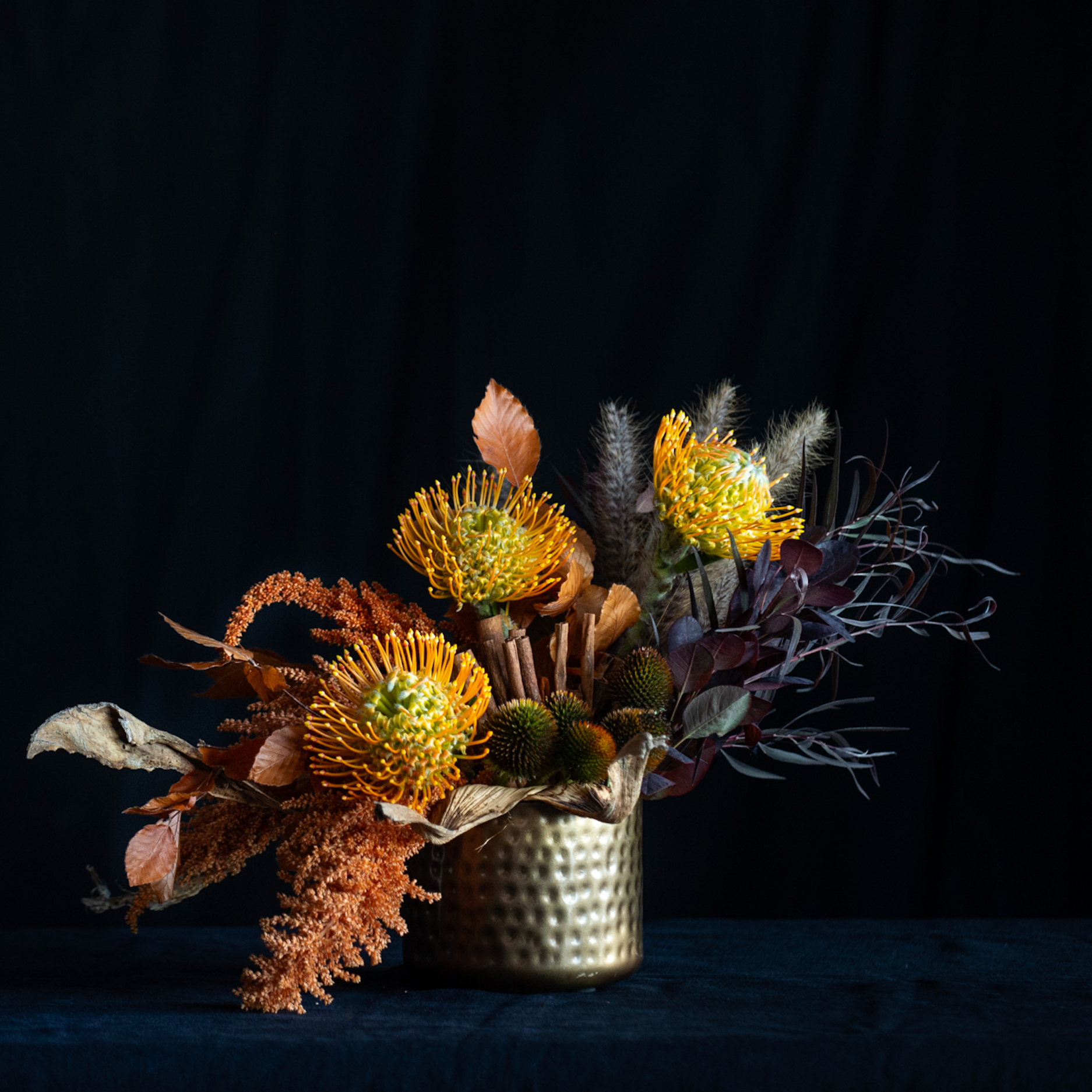 Fall floral arrangement with pincushion protea, amaranthus, pods, and autumn leaves in a hammered gold vase.
