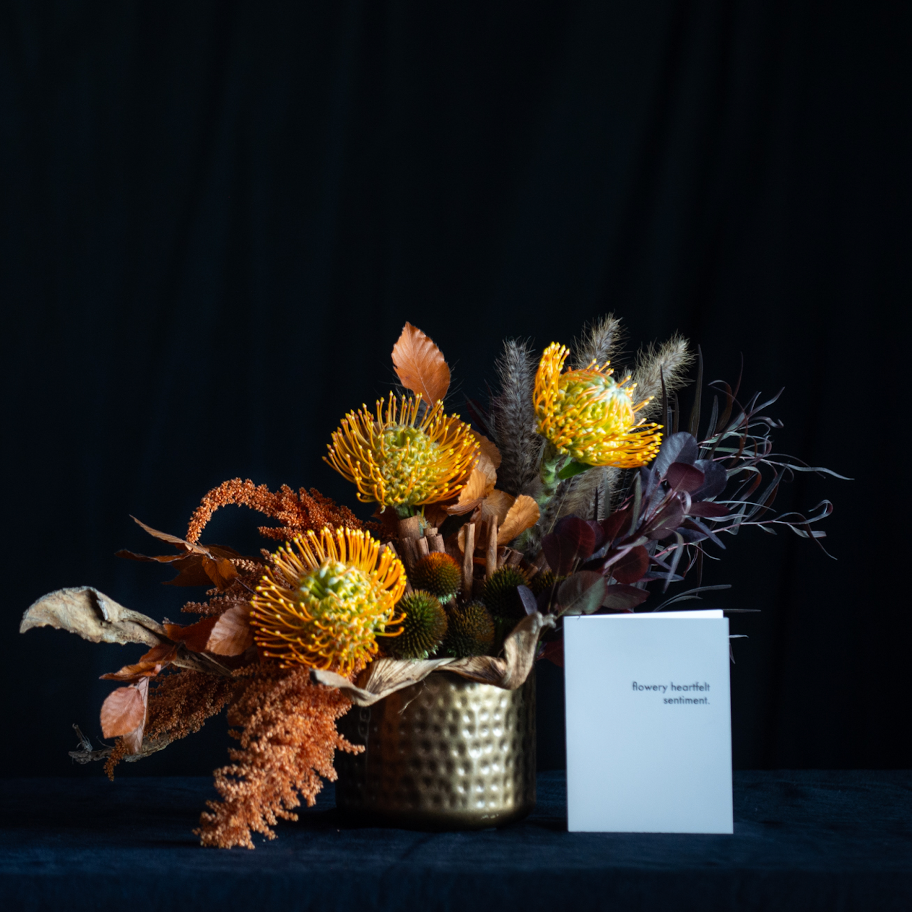 Fall floral arrangement with pincushion protea, amaranthus, pods, and autumn leaves in a hammered gold vase.