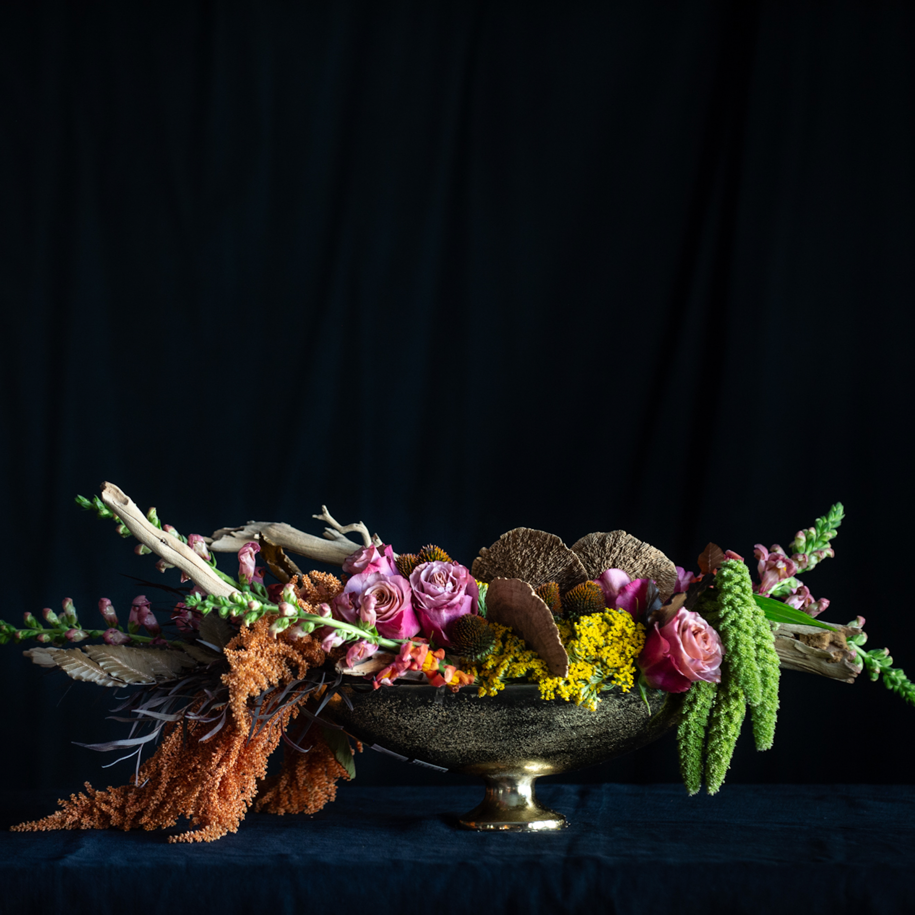 Autumn floral centerpiece in a bronze compote featuring roses, snapdragons, amaranthus, yellow yarrow, dried pods, and driftwood accents against a dark background.