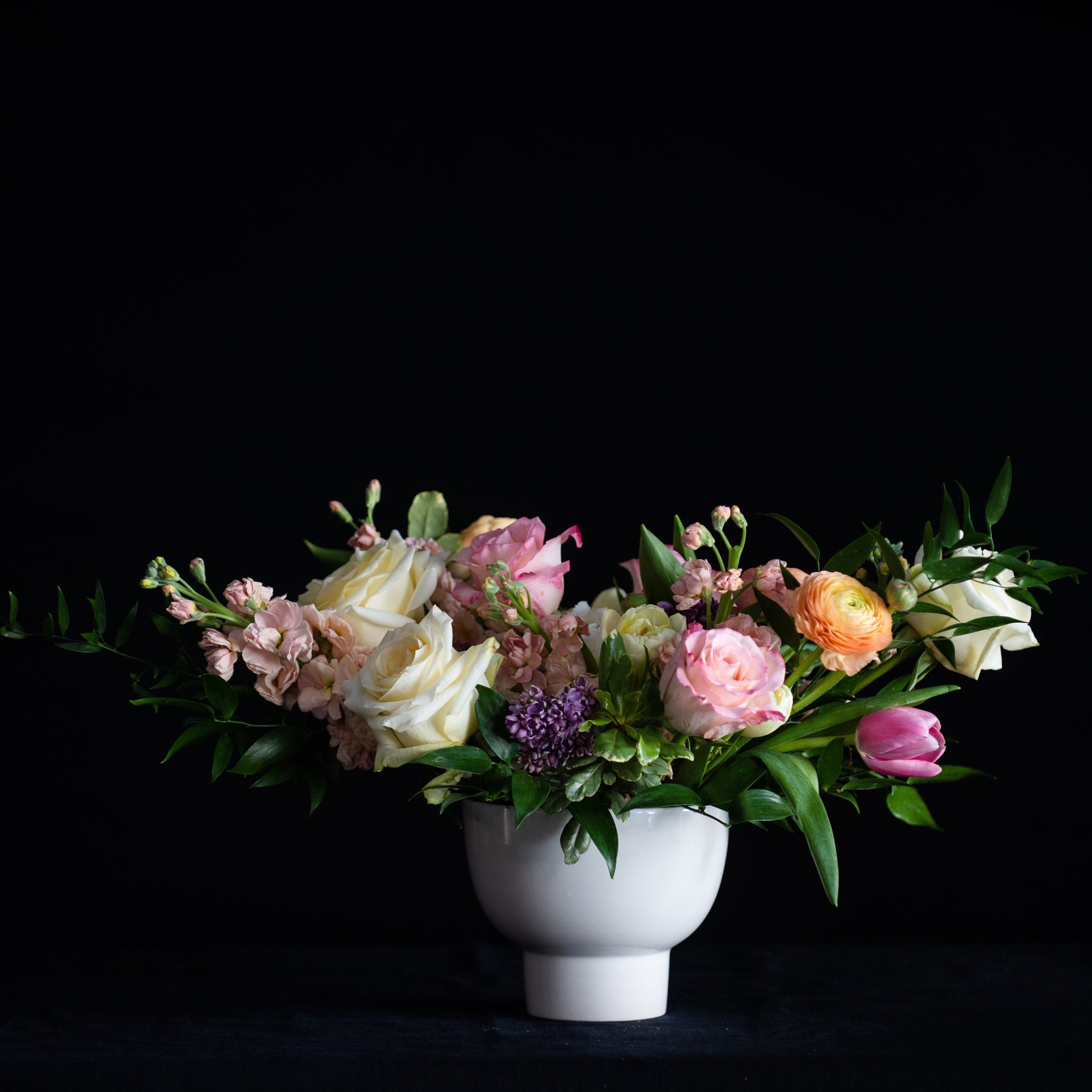 A white pedestal vase is filled with roses, tulips, lilac, stock, and ranunculus. 