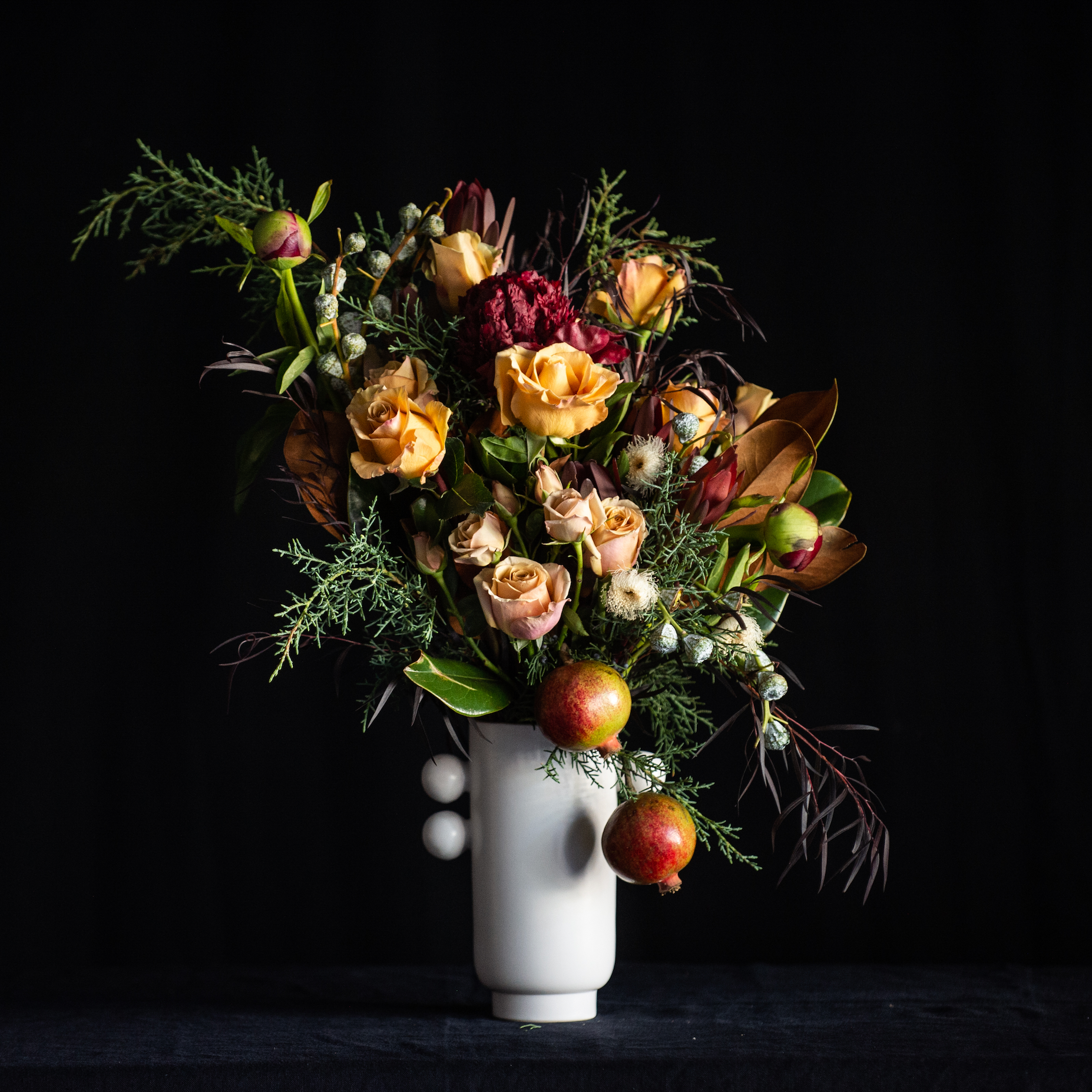 Design of a dozen golden roses with peonies and holiday greens with a pomegranate