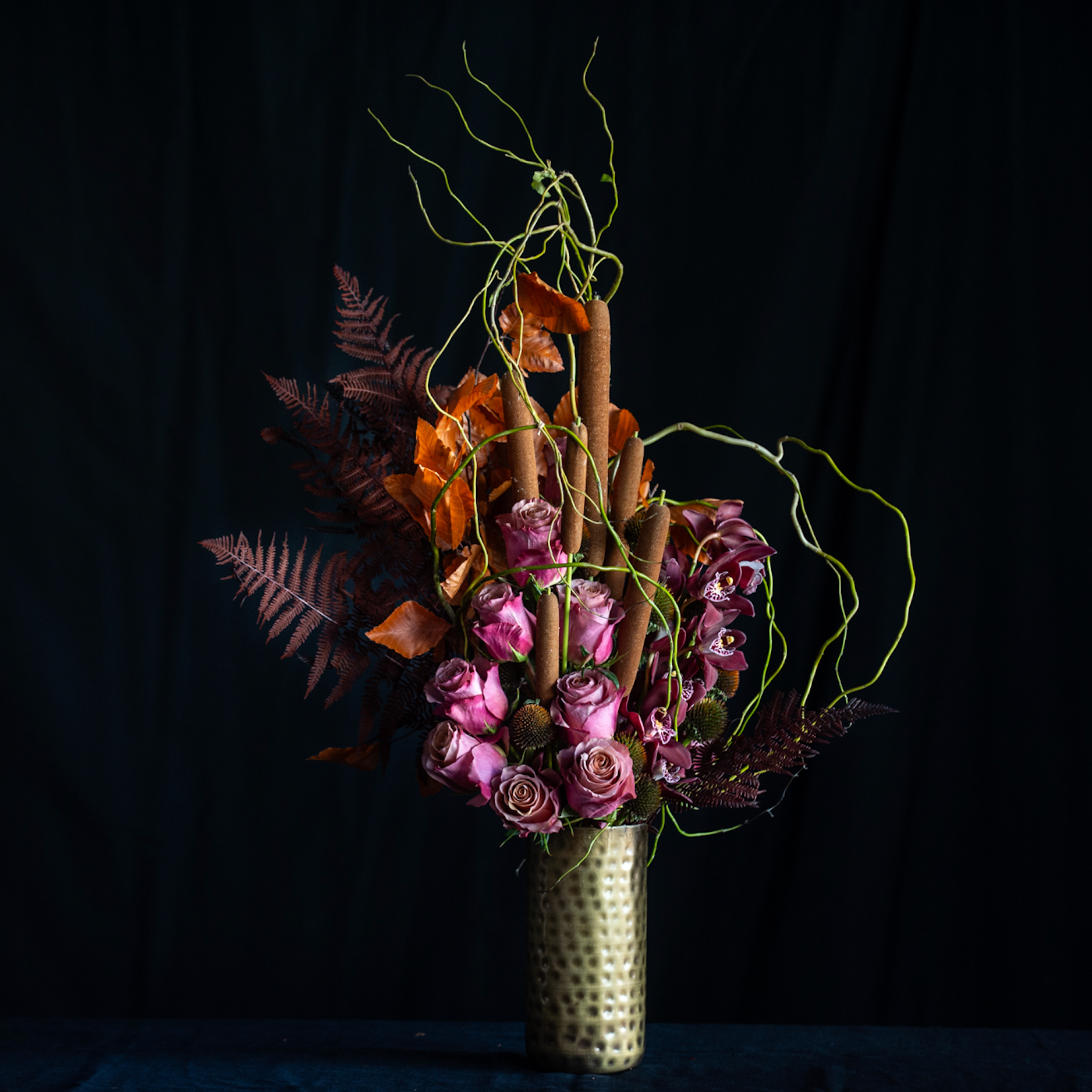 Fall floral arrangement with mauve roses, orchids, cattails, pods, and autumn foliage in a hammered gold vase.
