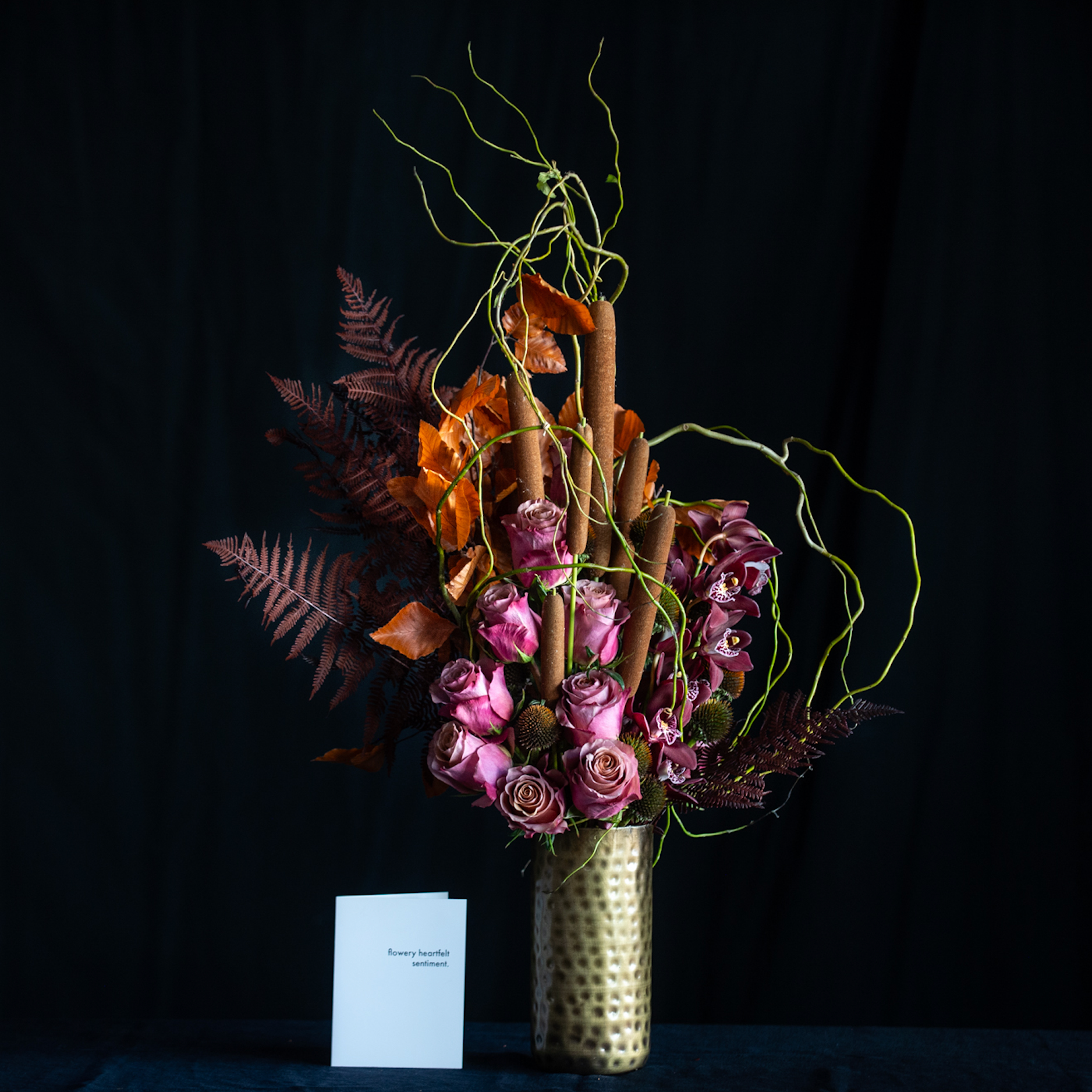 Fall floral arrangement with mauve roses, orchids, cattails, pods, and autumn foliage in a hammered gold vase.