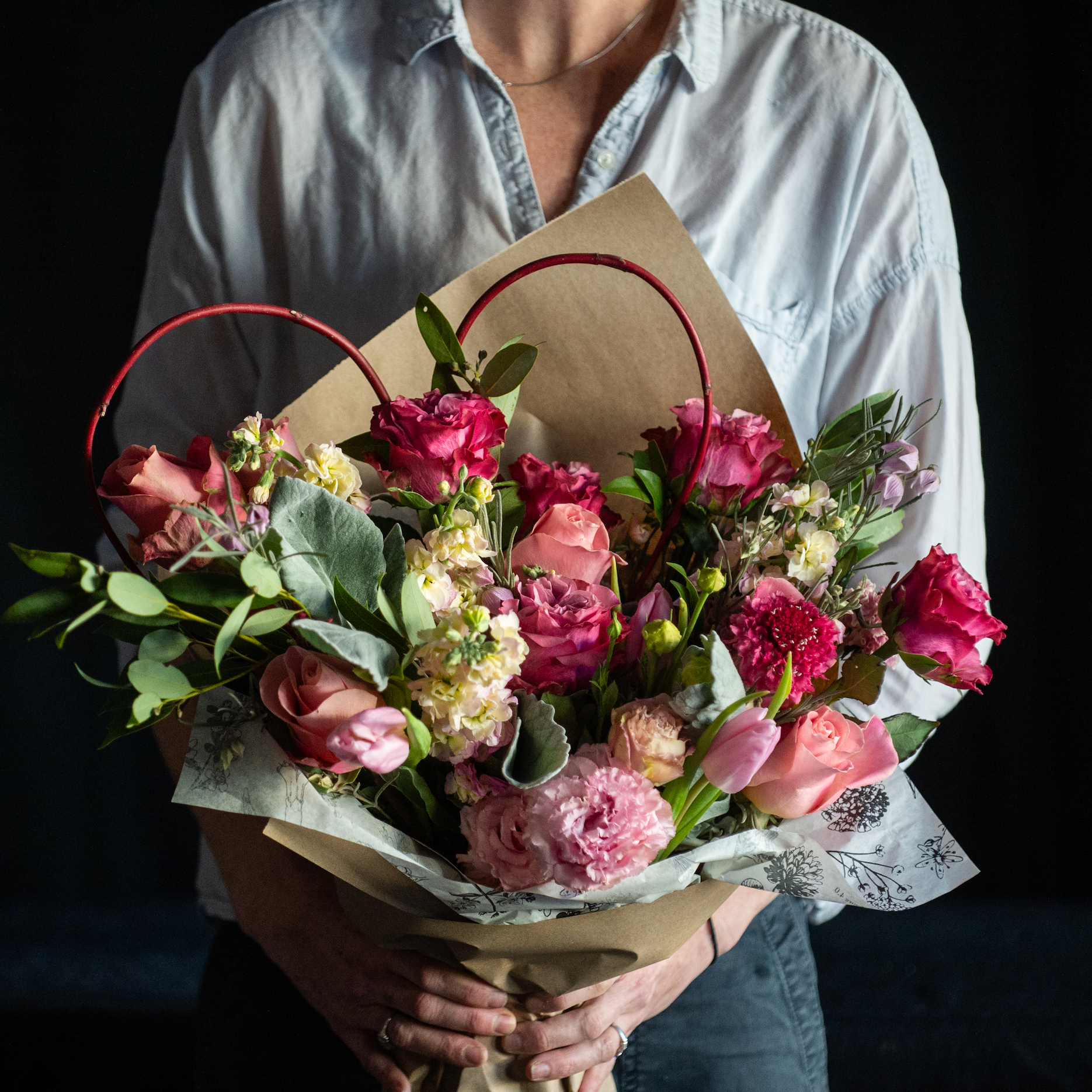 A wrapped bouquet of pink roses with premium accent stems with a red branch heart. 