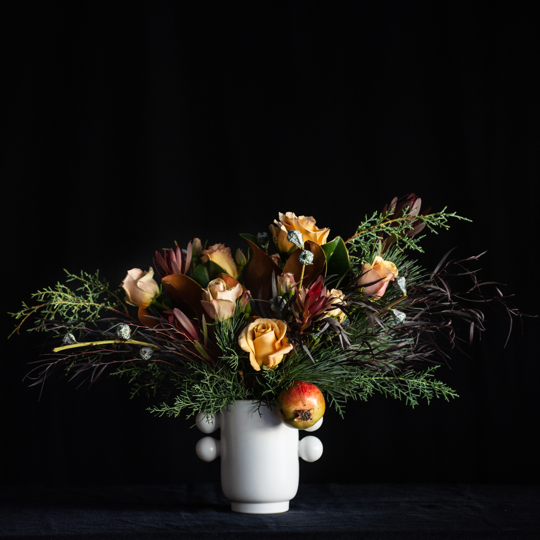 Holiday floral design with roses, pomegranate, and holiday greens in a festive white vase