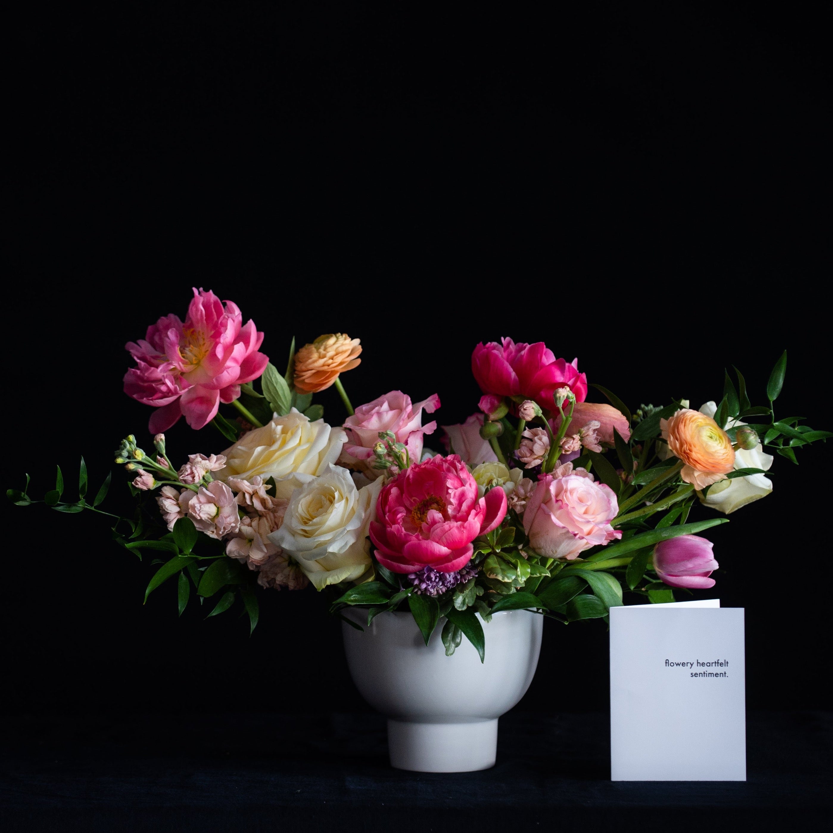 A modern pedestal vase overflows with pastel tulips, roses, stock, and ranunculus with option to add peonies.