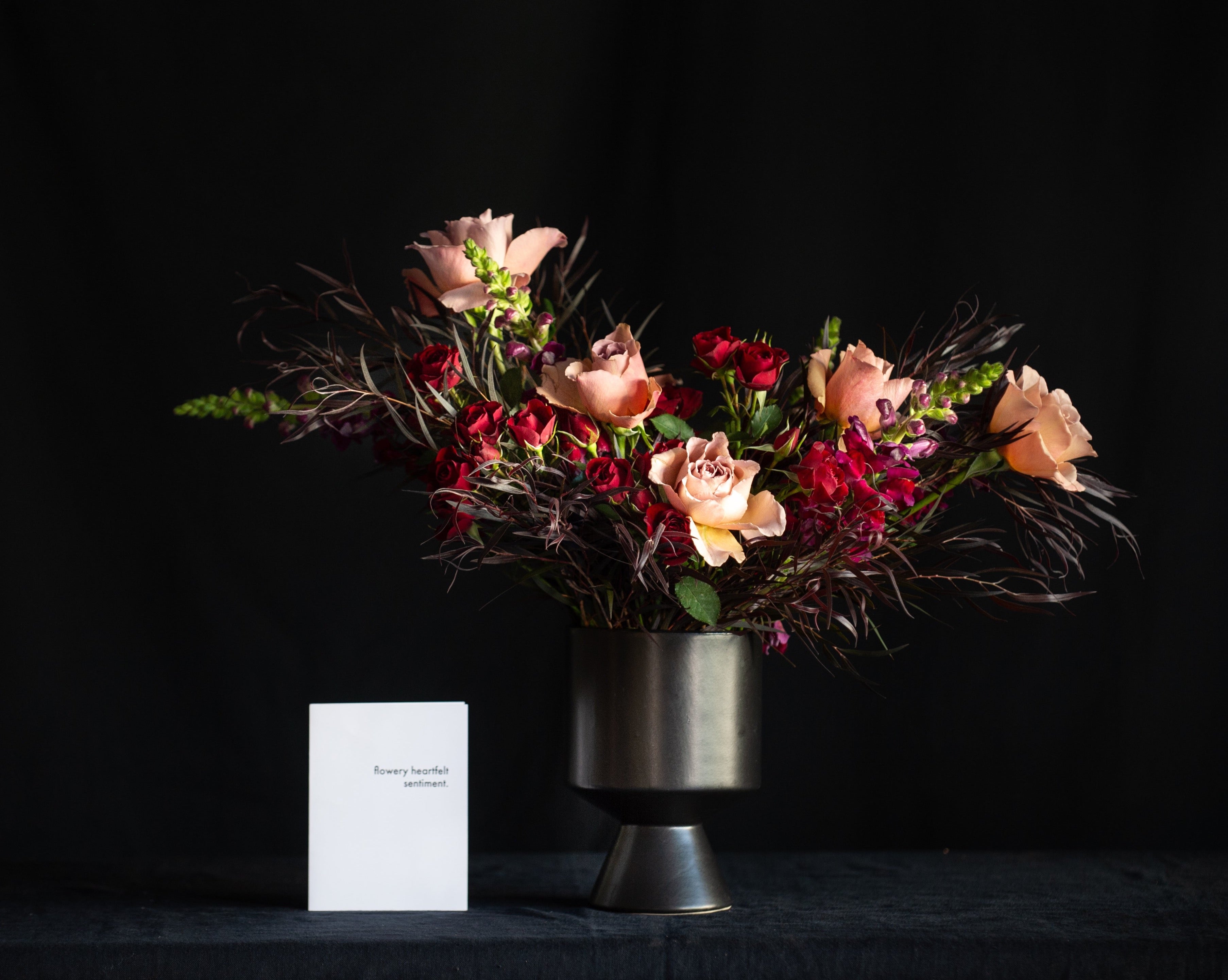 Garden roses, snap dragons, spray roses, and dark foliage in a black vase for valentines day