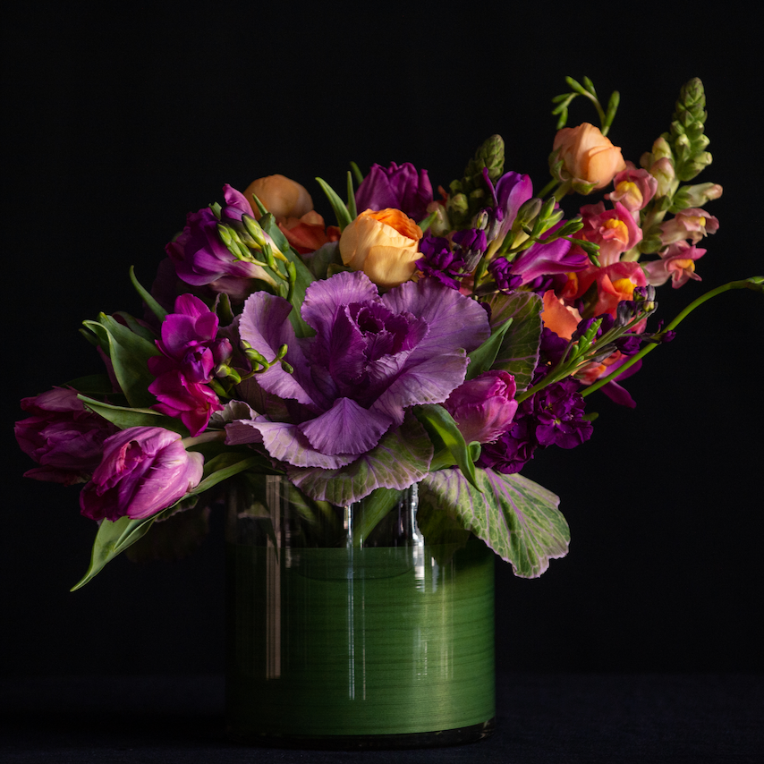 An asymmetrical design of kale, freesia, ranunculus, and snapdragons in a leaf lined container. Made by a local florist. 