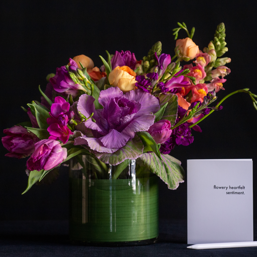 An asymmetrical design of kale, freesia, ranunculus, and snapdragons in a leaf lined container. Made by a local florist. 