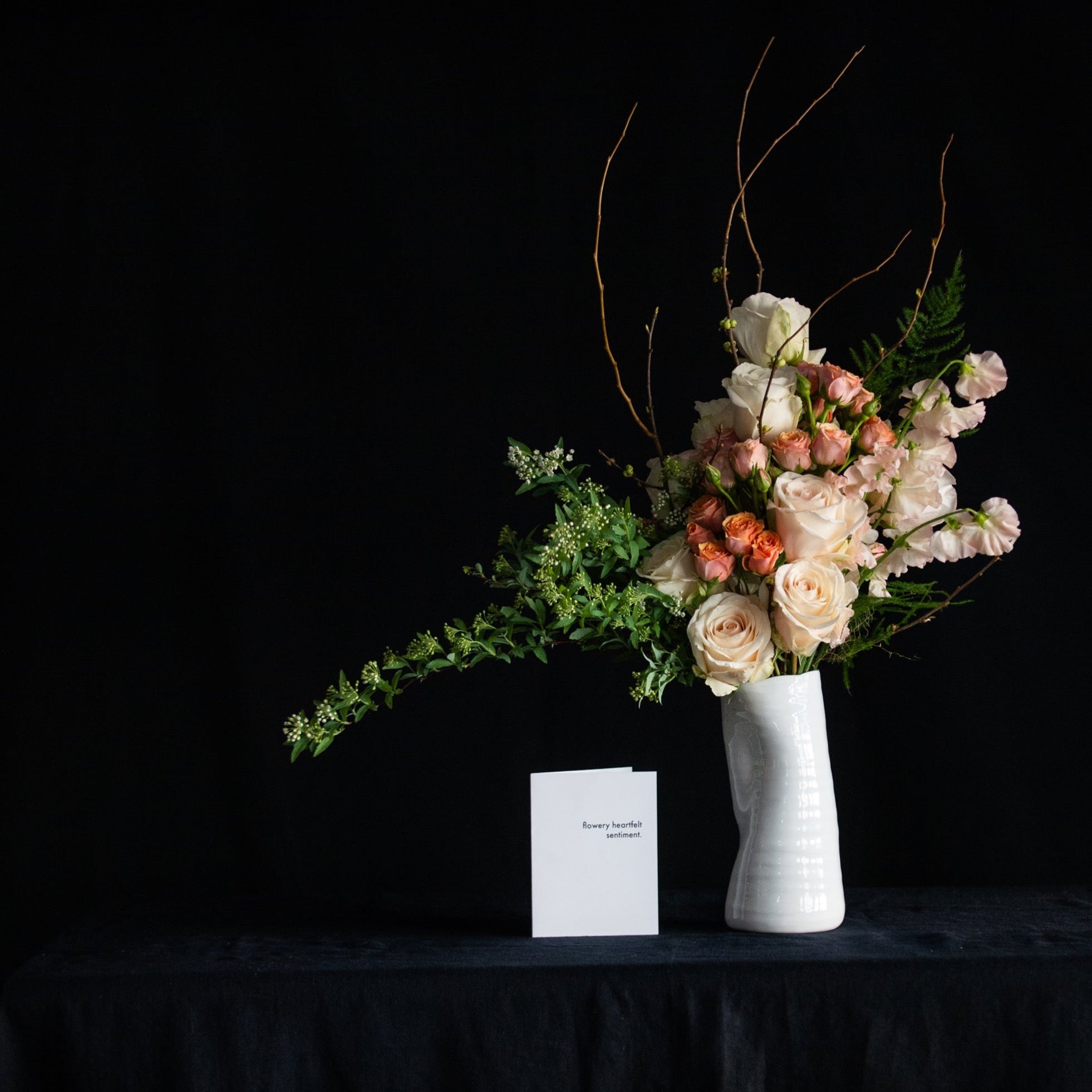 A dozen pastel roses with sweet peas, blooming branches and seasonal accents in a white ceramic vase. 