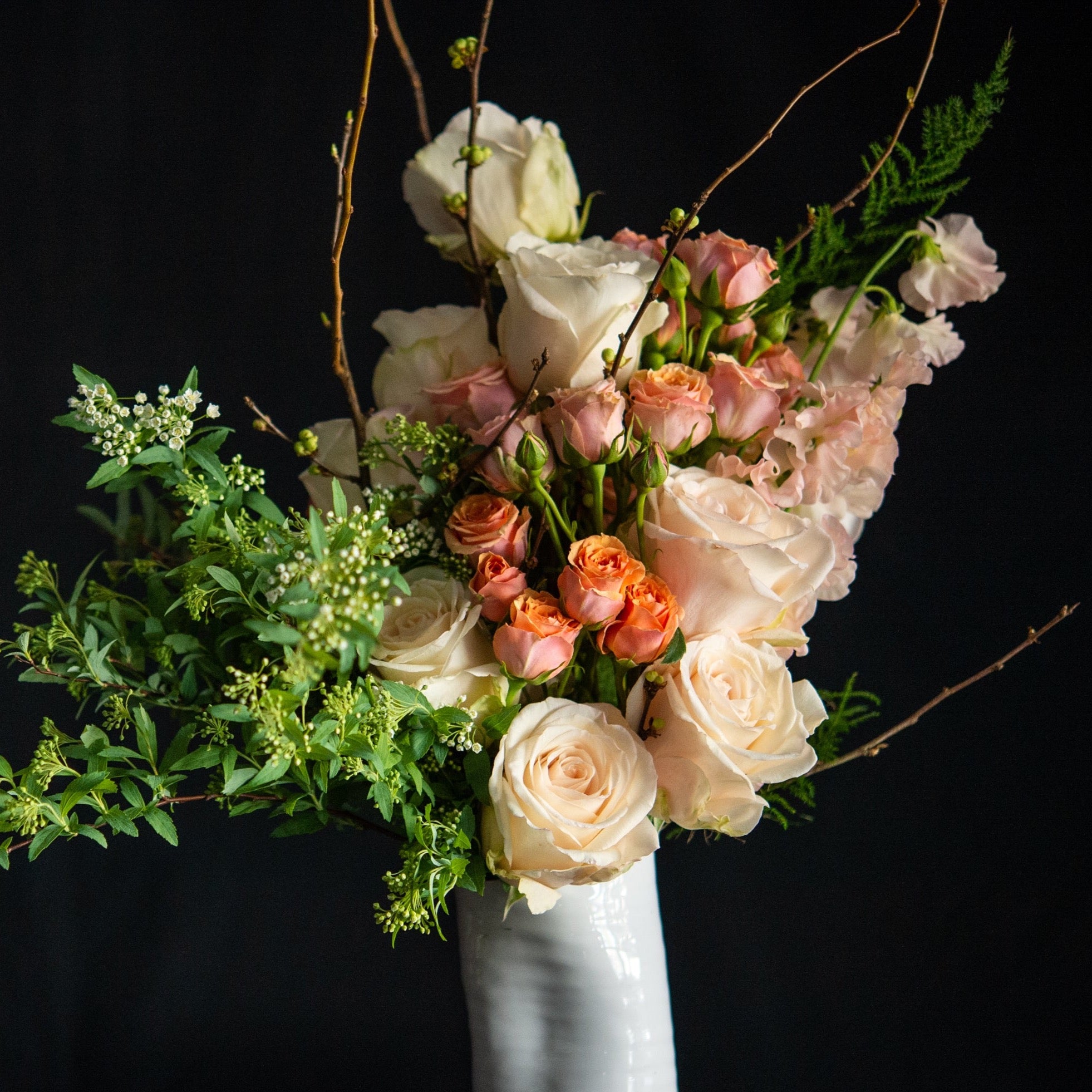 A dozen pastel roses with sweet peas, blooming branches and seasonal accents in a white ceramic vase. 