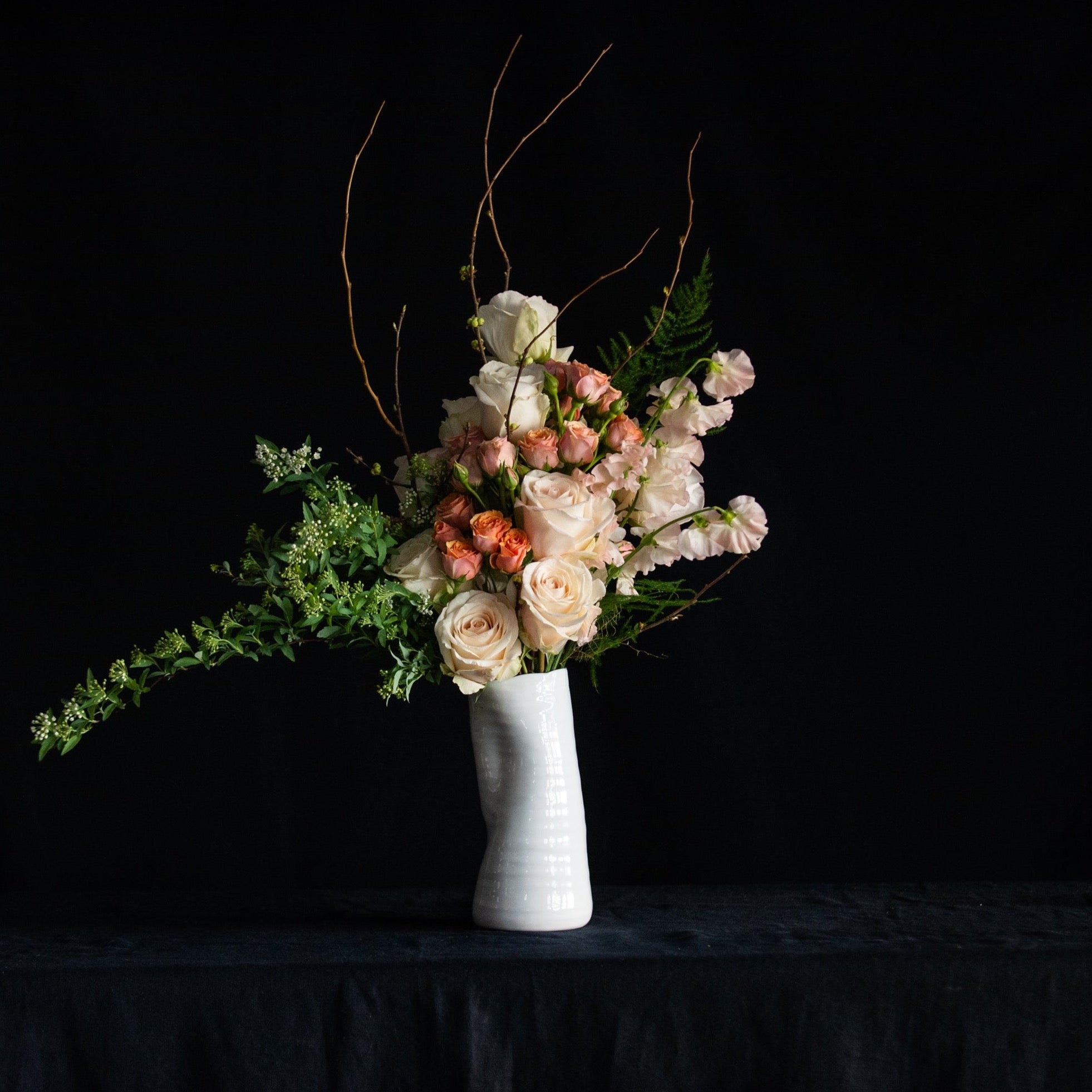 A dozen pastel roses with sweet peas, blooming branches and seasonal accents in a white ceramic vase. 