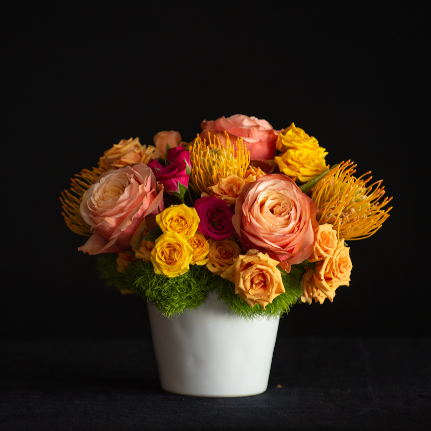 Lush compact summer design of roses and tropicals in sunset tones fill a white ceramic vase. Send flowers from a local florist!